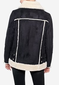 Kvinde iført en sort lammeskindsjakke med hvidt pelsdetalje på kraven, bagsider og kant, vist fra ryggen.