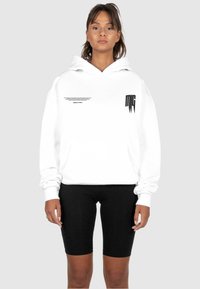 Weißer Kapuzenpullover mit einer Vorderasche, der schwarzen Text und ein Logo auf der Brust aufweist. Kombiniert mit schwarzen Radshorts.