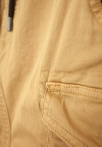 Kaki tkanina s glatkom teksturom, koja sadrži bočnu džepu i suptilne detalje šivanja, prikazana u bliskom pogledu.