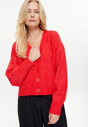 Rode gebreide cardigan met een V-hals, geribbelde textuur, oversized mouwen en grote knopen. Korte lengte gecombineerd met zwarte broeken.