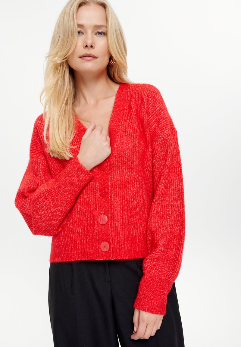 Cardigan rosso lavorato a maglia con scollatura a V, texture a coste, maniche oversize e grandi bottoni. Lunghezza corta abbinata a pantaloni neri.