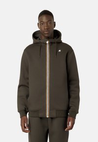 Felpa con zip di colore verde scuro, dotata di una cerniera frontale e caratterizzata da una striscia multicolore, cordini regolabili e un piccolo logo bianco sul petto.