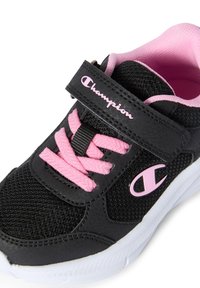 Schwarzer Sportschuh mit pinken Akzenten, ausgestattet mit einem Klettverschluss, strukturiertem Material und einer kontrastierenden weißen Sohle. Enthält pinke Schnürsenkel.
