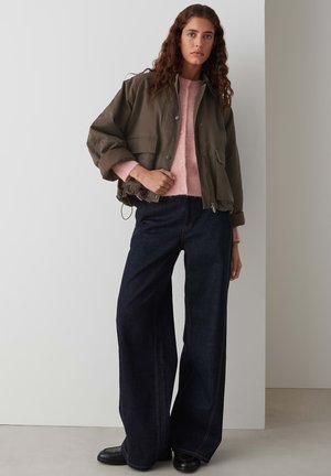 Khaki bomberjack met een oversized silhouet, gecombineerd met een lichtroze gebreide trui en donkere wijde jeans. Zwarte schoenen maken de look af.