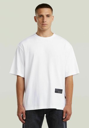 Młody mężczyzna stojący zwrócony przodem, ubrany w prosty biały oversize t-shirt i ciemne dżinsy na jasnoszarym tle.