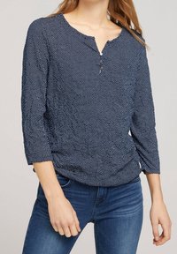 Marineblå langærmet bluse med et struktureret mønster af små hvide prikker, der har en knap lukning ved halsen og en afslappet pasform.