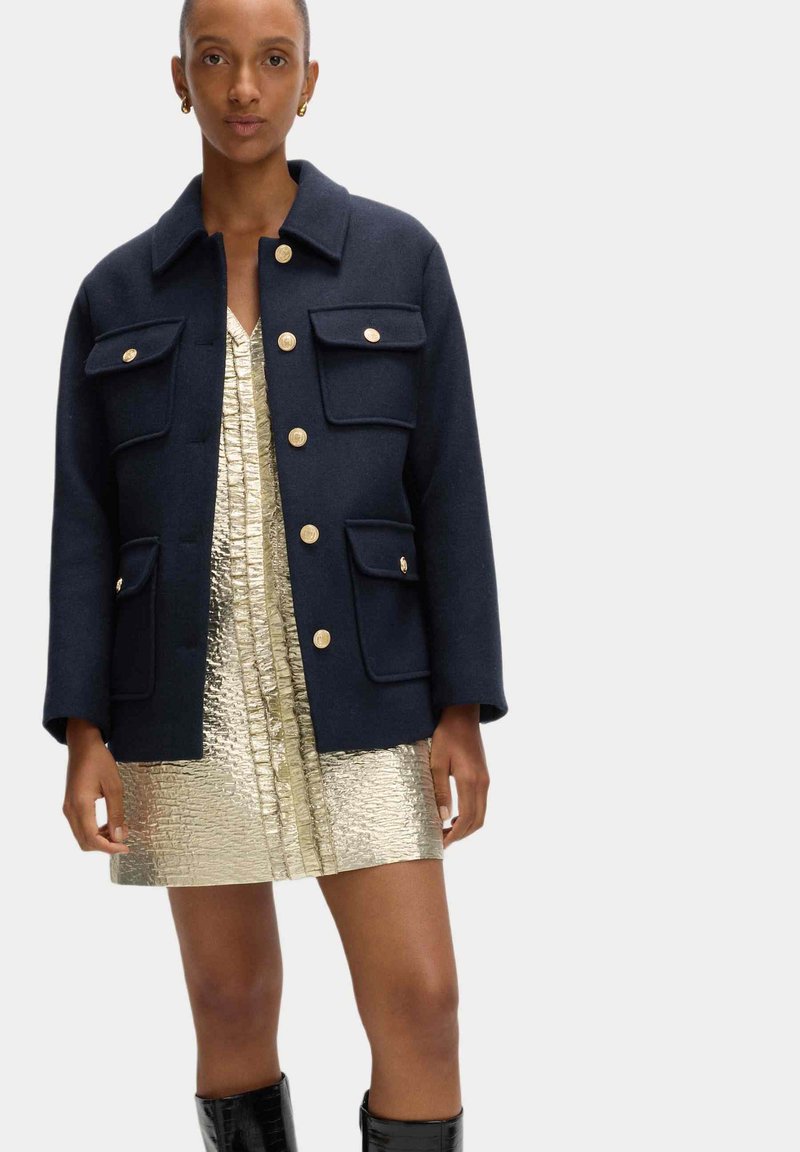 Marineblaues Wollmischjackett mit vier Vordertaschen und goldenen Knöpfen, getragen über einem metallisch goldenen Kleid mit strukturierter Oberfläche.