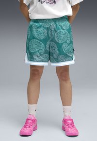 Persoon in teal paisley-gepatroneerde mesh short, witte sokken tot de kuit, en felroze sport schoenen, staande op een grijze achtergrond.