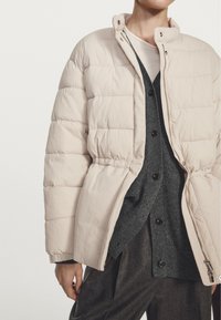 Persona che indossa un piumino beige sopra un cardigan grigio scuro con bottoni e pantaloni scuri, in piedi contro uno sfondo chiaro e uniforme.