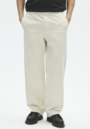 Mann trägt lockere, cremeweiße Hose, schwarze Schuhe und schwarzes Hemd, steht mit den Händen in den Taschen vor einem schlichten Hintergrund.