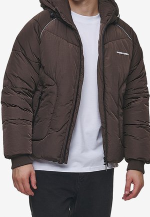 Braune Steppjacke mit gefütterter Kapuze, Frontreißverschluss, Seitentaschen und kontrastierendem weißen Nähten. Getragen über einem weißen Shirt.