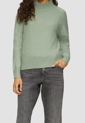 Frau trägt einen hellgrünen, gerippten Strickpullover mit langen Ärmeln und ausgewaschenen schwarzen Jeans, steht mit einer Hand in der Tasche.