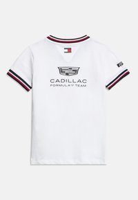 Λευκό κοντομάνικο μπλουζάκι με το λογότυπο της ομάδας Cadillac Formula 1 στην πλάτη, κόκκινος, λευκός και μπλε ριγωτός γιακάς και μανσέτες, ετικέτα Tommy Hilfiger.