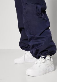 Witte Nike Air Force 1 sneakers van textuurleder met perforaties, gecombineerd met losse marineblauwe nylon cargobroeken.