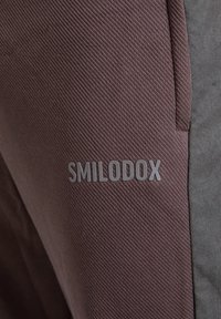 Bruine joggers van een geweven stof met een gladde grijze bies, met een opvallend "SMILODOX" logo in lichtgrijs op de dij.