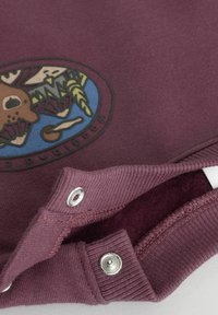 Mørkerød sweatshirt med rundt logo, der viser en bjørn, planter og en blå baggrund. Inkluderer sølvfarvede trykknapper ved kraven.