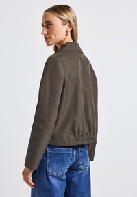 Brun, lättviktsjacka med krage och elastisk midja. Har långa ärmar och en slät textur, kombinerad med blå jeans.
