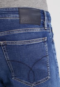 Jeans de mezclilla de color azul oscuro con una etiqueta de cuero de Calvin Klein Jeans en la cintura y costuras detalladas en los bolsillos traseros.