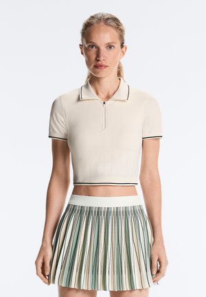 Femeie care poartă un polo crem cu mâneci scurte cropped și o fustă plisată cu dungi verticale verzi, crem și negre.