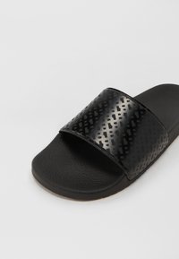 Sort slidesandal med en tekstureret rem, der har et geometrisk mønster. Sålen er lavet af gummi med en tekstureret overflade for bedre greb.