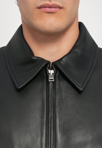 Veste en cuir noir avec un large col, fermeture zippée centrale et texture lisse, mettant en valeur le design élégant et le savoir-faire.