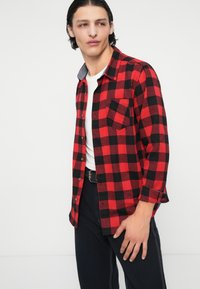 Camicia di flanella a quadri rossi e neri, con colletto grigio, tasca frontale singola e chiusura con bottoni, abbinata a pantaloni neri.