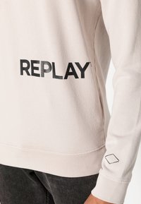 Ljus beige sweatshirt med svart "REPLAY"-text, ribbade muddar och en accentuerad diamantform på ärmen, tillverkad av mjukt tyg.