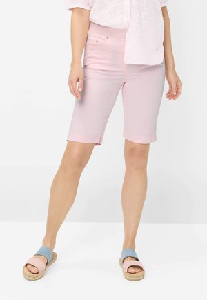 Kvinde iført lyserøde knælange shorts, en pink top og pink og blå slip-on sandaler, stående mod en ensfarvet hvid baggrund.