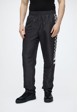 Mężczyzna w czarnych spodniach sportowych z białym, kratkowanym i czaszkowym wzorem po bokach, w czarnych trampkach, stojący na białym tle.