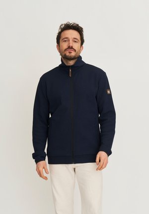 Man draagt een marineblauwe trui met rits en hoge kraag en witte broek, staand tegen een effen lichte achtergrond.