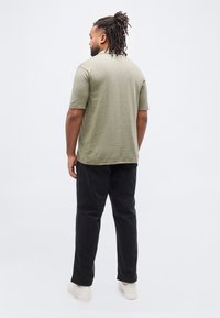 Άνδρας με ντραντλοκς που φοράει ελιά πράσινο κοντομάνικο πουκάμισο, μαύρο παντελόνι και λευκά αθλητικά παπούτσια, στέκεται με την πλάτη γυρισμένη σε λευκό φόντο.