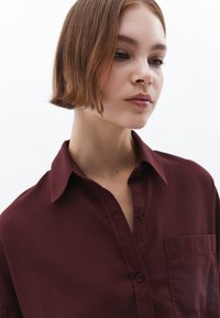 Camicia bordeaux con colletto classico, chiusura frontale con bottoni e taschino sul petto. Il tessuto appare liscio con una texture opaca.