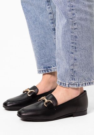 Mocassins en cuir noir avec détail mors doré portés avec un jean court bleu clair pieds nus.