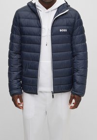 Muž v námořnickém prošívaném kabátu s logem "BOSS" přes bílou mikinu a bílými kalhotami, stojící s uvolněnýma rukama podél těla.