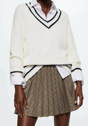 Femme portant un pull à col en V blanc avec des rayures noires sur une chemise blanche et une jupe plissée à carreaux marron.