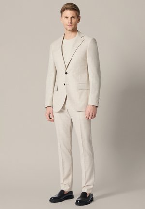 Mężczyzna w jasnobeżowym garniturze z jednorzędową marynarką na jeden guzik, dopasowanymi spodniami, białą koszulą i czarnymi mokasynami, stojący na beżowym tle.