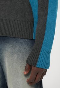Nahaufnahme einer Hand, die an der Seite ruht, bekleidet mit einem blauen und dunkelgrauen Strickpullover mit gerippten Bündchen und ausgewaschenen blauen Jeans.