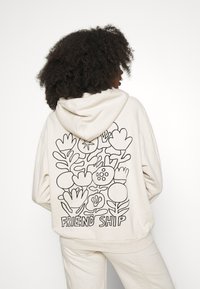Personne aux cheveux bouclés portant un sweat à capuche crème orné d'une illustration florale noire en ligne et du mot « AMITIÉ » dans le dos, sur fond blanc.