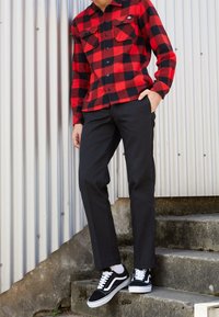 Camisa de franela de cuadros roja y negra con bolsillos abotonados, combinada con pantalones negros y zapatillas negras y blancas. Escenario al aire libre con un fondo metálico.