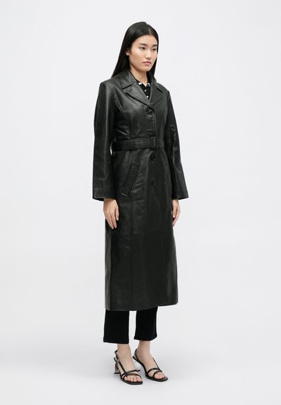 Cappotto trench in pelle nera con vita cinturata, chiusura a bottoni, due tasche frontali e texture liscia, indossato su pantaloni neri a sigaretta.