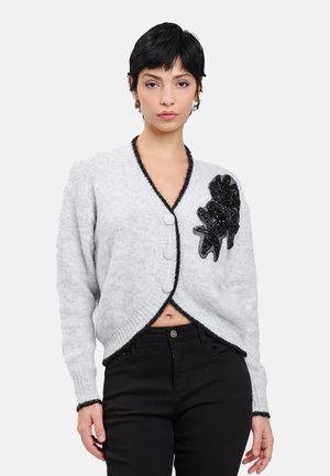 Donna con capelli corti neri indossa un cardigan grigio con rifiniture nere e un'applicazione floreale nera, abbinato a pantaloni neri a vita alta.