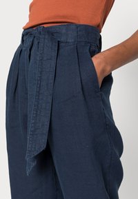 Pantalones de lino azul marino con cintura alta, frente plisado y cinturón atado. Presenta bolsillos laterales y una tela texturizada.