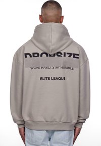 Sweat à capuche beige avec un grand logo noir « DROPSIZE » dans le dos, accompagné des inscriptions « WORK HARD, STAY HUMBLE » et « ELITE LEAGUE » en plus petit.