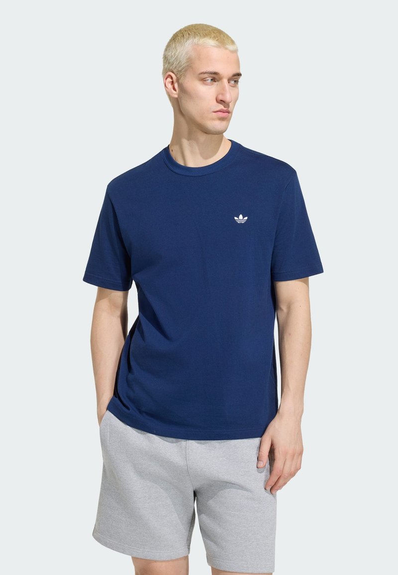 Jeune homme aux cheveux blonds courts portant un t-shirt Adidas bleu marine et un short gris clair, les mains dans les poches, regardant vers la droite.