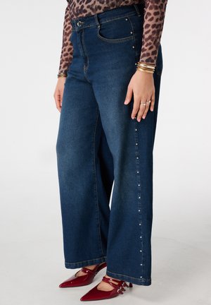 Femme portant un jean large bleu foncé avec des clous sur le côté, un haut imprimé léopard, des bracelets dorés et des talons pointus rouges, debout devant un fond blanc.