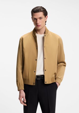 Mann trägt eine beige, geknöpfte Bomberjacke über einem weißen Hemd mit schwarzen Hosen und steht mit einer Hand in der Tasche vor weißem Hintergrund.