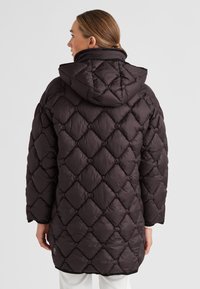 Frau in einem dunklen, gesteppte, gefütterten Kapuzenmantel, von hinten gezeigt, mit zu einem tiefen Dutt gebundenem Haar vor einem schlichten, hellen Hintergrund.