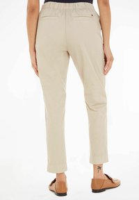 Pantaloni beige con vita elastica e gambe affusolate, dotati di due tasche posteriori e un dettaglio logo discreto. Indossati con scarpe marroni slip-on.