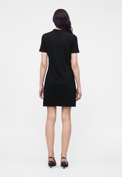 Femme aux longs cheveux foncés portant une robe noire à manches courtes et des chaussures à talons noires, debout dos à la caméra.