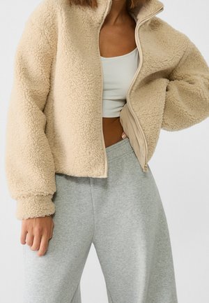 Vrouw draagt een beige sherpa jas met rits over een wit cropped top en lichtgrijze joggingbroek met één hand in de zak.